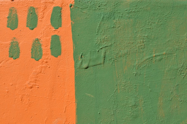 A textured wall is painted in two contrasting colors: a vibrant orange on the left and a muted green on the right. The orange section includes six oval green paint marks arranged in two vertical columns.