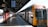 A train is stationed at a platform under a covered structure. The platform is empty, and a timetable board hangs overhead displaying train times and stops. The train has a yellow and silver exterior marked with 'Intercity'. Sunlight casts shadows across the tiled floor.