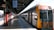 A train is stationed at a platform under a covered structure. The platform is empty, and a timetable board hangs overhead displaying train times and stops. The train has a yellow and silver exterior marked with 'Intercity'. Sunlight casts shadows across the tiled floor.