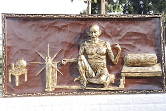 A relief sculpture depicts a seated figure engaged in spinning at a traditional wheel. The figure has an emaciated appearance, wearing simple cloth wrapped around. On the left, there's a pot on a stool, and on the right, a rolled-up mattress is placed on a bench.
