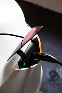 A close-up view of an electric vehicle charging port with the charger plugged in. The car appears to be white, and the charging cable has a visible label with a company name. The tail light is partially visible with a red lens and a sleek design. The scene suggests a modern and clean aesthetic typical of electric vehicles.