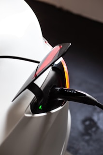 A close-up view of an electric vehicle charging port with the charger plugged in. The car appears to be white, and the charging cable has a visible label with a company name. The tail light is partially visible with a red lens and a sleek design. The scene suggests a modern and clean aesthetic typical of electric vehicles.