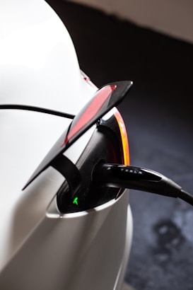 A close-up view of an electric vehicle charging port with the charger plugged in. The car appears to be white, and the charging cable has a visible label with a company name. The tail light is partially visible with a red lens and a sleek design. The scene suggests a modern and clean aesthetic typical of electric vehicles.