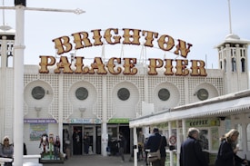 A large sign with bold, ornate lettering reading 'Brighton Palace Pier' is prominently displayed on a historic-looking building with a classic seaside architecture featuring arched windows. People are walking around the entrance, some headed towards the arcade with various attractions like a carousel and coffee shop signage visible.