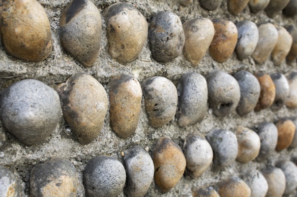 A close-up view of a textured wall constructed with smooth, rounded stones embedded in a concrete-like surface. The stones vary in color from gray to brown and are arranged in a uniform pattern.