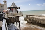 A seaside pier extends over the ocean with a small pavilion decorated with merchandise, including clothing and souvenirs. The background features a calm sea and a stone breakwater with a solitary walker. The sky is partly cloudy.