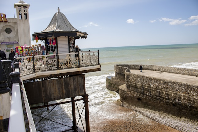 A seaside pier extends over the ocean with a small pavilion decorated with merchandise, including clothing and souvenirs. The background features a calm sea and a stone breakwater with a solitary walker. The sky is partly cloudy.