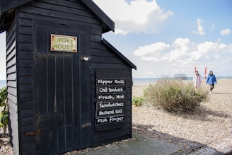 A small black wooden smokehouse is situated on a pebble beach. It features a sign listing food items such as kipper roll, crab sandwich, and fish finger. Two people are walking along the beach in the background under a partly cloudy sky.