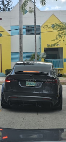 A dark-colored Tesla is parked on a street in front of a building with yellow and white geometric shapes and blue accents. There are palm trees in the background, and the car's license plate is visible.