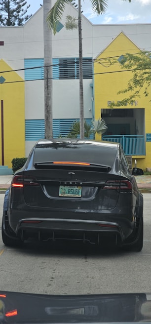 A dark-colored Tesla is parked on a street in front of a building with yellow and white geometric shapes and blue accents. There are palm trees in the background, and the car's license plate is visible.