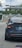 A black Tesla car is at a traffic light intersection, visible from the rear. The vehicle has a Florida license plate, and the traffic light is green. Another car is in front of the Tesla. In the background, there are trees, stairs leading up to a building, and a cloudy sky with a palm tree on the left.