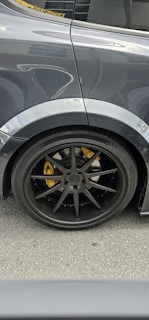 The image features the rear side of a car, focusing on the wheel and tire. The car has a dark, glossy finish with a black wheel rim and a visible yellow brake caliper. Reflections and details of the car's surface are noticeable, with part of the window and a performance sticker visible.
