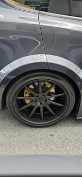 The image features the rear side of a car, focusing on the wheel and tire. The car has a dark, glossy finish with a black wheel rim and a visible yellow brake caliper. Reflections and details of the car's surface are noticeable, with part of the window and a performance sticker visible.