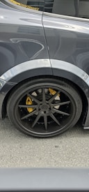 The image features the rear side of a car, focusing on the wheel and tire. The car has a dark, glossy finish with a black wheel rim and a visible yellow brake caliper. Reflections and details of the car's surface are noticeable, with part of the window and a performance sticker visible.