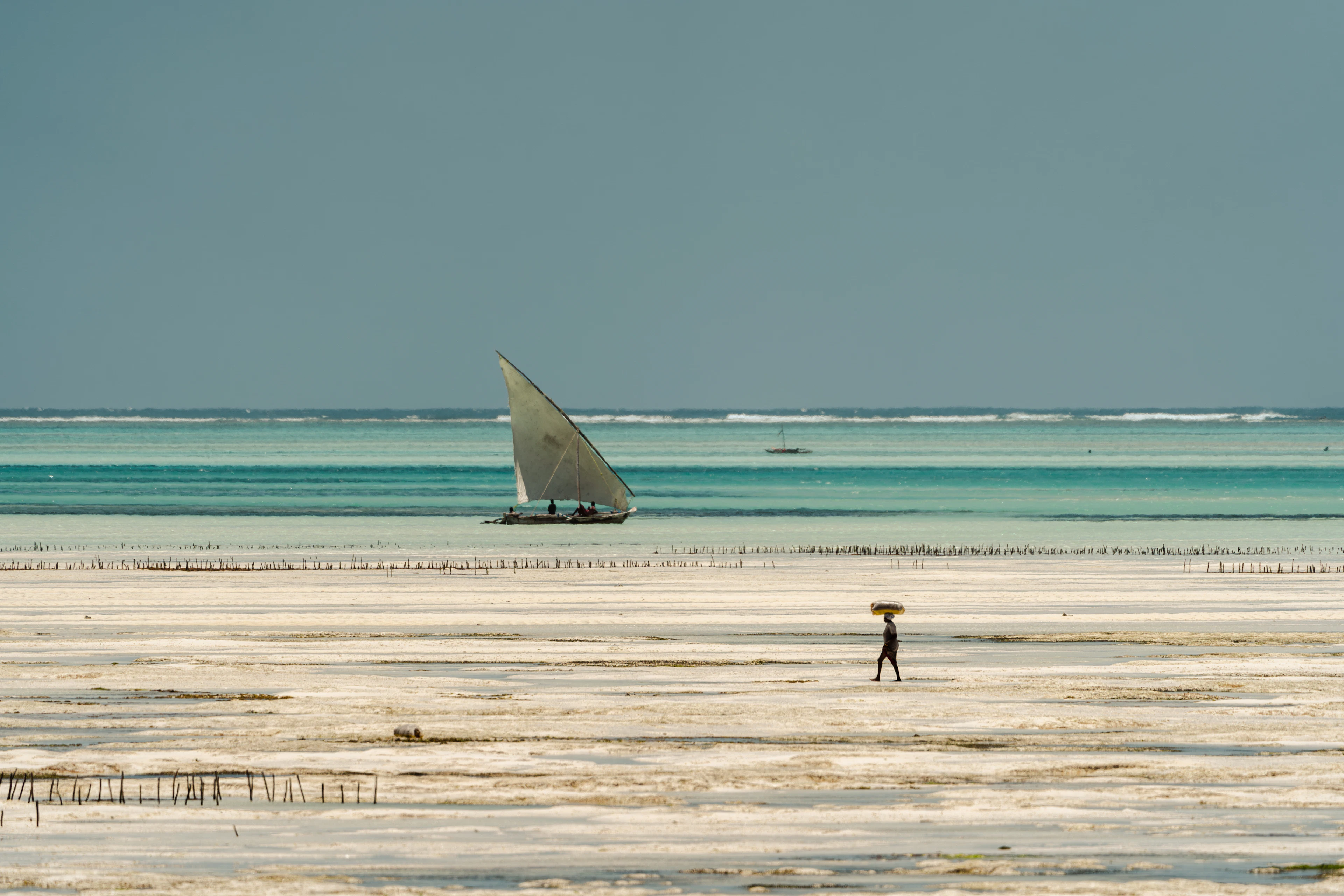 Low-tide flats at Jambiani, Zanzibar, with a woman carrying a load on her head and a dhow sailing beyond