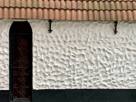 A textured white stone wall with a dark, narrow arched window featuring intricate metalwork. The building is topped with a terracotta tiled roof, and there are two metal fixtures attached to the wall for support.