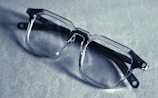 A pair of transparent, rectangular eyeglasses rests on a textured surface, casting a shadow. The glasses have thick frames and prominent hinges, suggesting a bold, modern style. The lighting creates a soft, muted tone across the image.