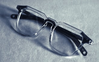 A pair of transparent, rectangular eyeglasses rests on a textured surface, casting a shadow. The glasses have thick frames and prominent hinges, suggesting a bold, modern style. The lighting creates a soft, muted tone across the image.