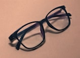 A pair of black, large-framed eyeglasses rests on a plain, light brown surface. The glasses have a sleek design, and their lenses slightly reflect the ambient light. Shadows from the glasses fall softly to the side, adding depth to the composition.