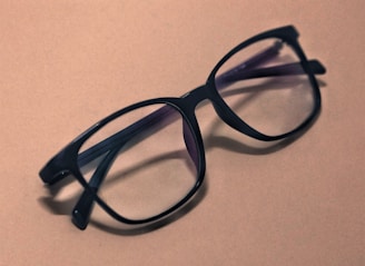 A pair of black, large-framed eyeglasses rests on a plain, light brown surface. The glasses have a sleek design, and their lenses slightly reflect the ambient light. Shadows from the glasses fall softly to the side, adding depth to the composition.