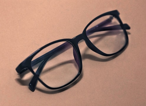 A pair of black, large-framed eyeglasses rests on a plain, light brown surface. The glasses have a sleek design, and their lenses slightly reflect the ambient light. Shadows from the glasses fall softly to the side, adding depth to the composition.