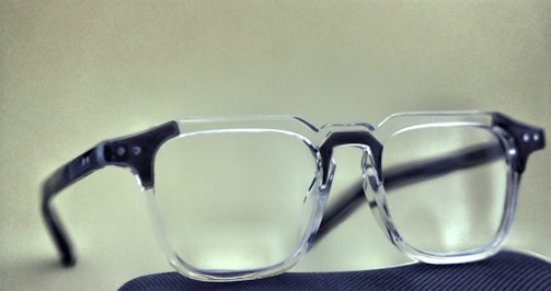 A pair of clear-framed eyeglasses with black temples rests on a dark textured surface. The background is a smooth beige color, contrasting with the transparency of the lenses and the solidity of the frame.