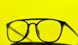 A pair of black-framed eyeglasses is placed against a bright yellow background. The glasses are centered, emphasizing their sleek design.