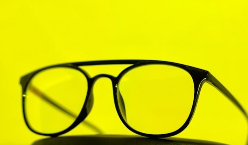 A pair of black-framed eyeglasses is placed against a bright yellow background. The glasses are centered, emphasizing their sleek design.