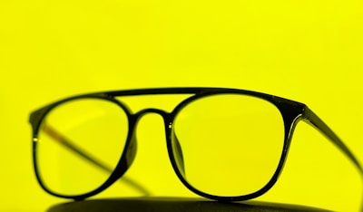 A pair of black-framed eyeglasses is placed against a bright yellow background. The glasses are centered, emphasizing their sleek design.