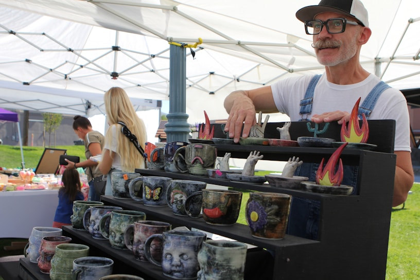 A vendor with glasses and a mustache displays colorful and artistic pottery cups on a black shelf in a market setting. The pottery features designs with hands, flames, and various motifs. In the background, people, including a woman with long blonde hair and a child, browse other market stalls under white tents.