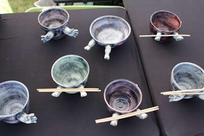 Several ceramic bowls with unique designs featuring sculpted arms and legs on a black surface are arranged in a scattered pattern. Each bowl has a pair of chopsticks resting on top. The bowls have a variety of colors, including hints of blue, purple, and red.