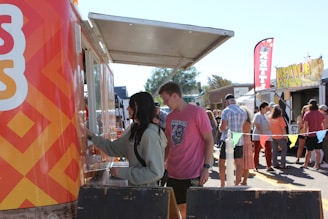 A bustling outdoor scene at a food market, featuring a brightly decorated food truck. Several people are queuing at the window to order. The crowd includes individuals in casual attire mingling around other food stalls in the background. The setting suggests a lively, community-oriented event under clear skies.