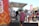 A bustling outdoor scene at a food market, featuring a brightly decorated food truck. Several people are queuing at the window to order. The crowd includes individuals in casual attire mingling around other food stalls in the background. The setting suggests a lively, community-oriented event under clear skies.