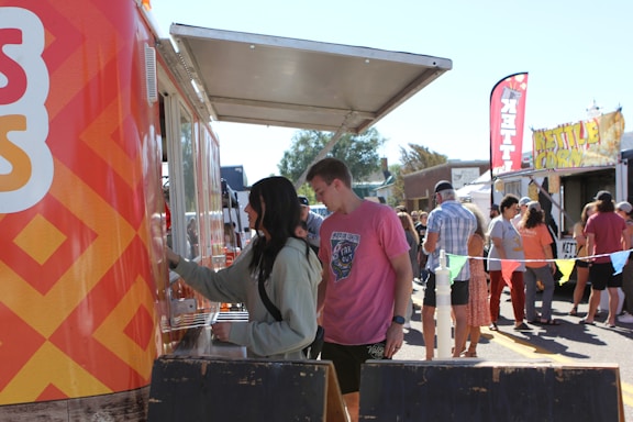 A bustling outdoor scene at a food market, featuring a brightly decorated food truck. Several people are queuing at the window to order. The crowd includes individuals in casual attire mingling around other food stalls in the background. The setting suggests a lively, community-oriented event under clear skies.
