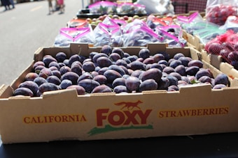 A cardboard box labeled as California strawberries is filled with fresh, ripe plums. Several other boxes and plastic bags containing fruits are visible on a table at an outdoor market. In the background, there are blurred figures of people walking on a sunny day.