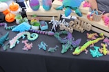 A display table is filled with colorful 3D-printed objects, including dragons, lizards, and a turtle. The items are multi-colored with shades of purple, teal, pink, and yellow. There are also carved wooden names and phrases and potted succulents. Additionally, Halloween-themed pumpkin decorations are visible, mingling with crafting tools on the side.