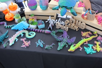 A display table is filled with colorful 3D-printed objects, including dragons, lizards, and a turtle. The items are multi-colored with shades of purple, teal, pink, and yellow. There are also carved wooden names and phrases and potted succulents. Additionally, Halloween-themed pumpkin decorations are visible, mingling with crafting tools on the side.
