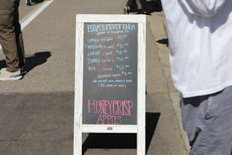 A wooden sidewalk sign with chalk writing advertises Isom's Fruit Farm products, listing the sizes and prices of different quantities of apples. The sign is framed in white and stands on a paved street with people walking nearby.