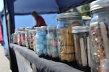 Several glass jars filled with various colorful sweets and cookies are arranged on a table covered with a dark cloth. The jars contain items like blue and pink foamy candies, multicolored candies, and cookies. In the background, there is a canopy tent and an out-of-focus figure that appears to be attending the setup.