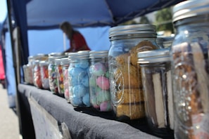 Several glass jars filled with various colorful sweets and cookies are arranged on a table covered with a dark cloth. The jars contain items like blue and pink foamy candies, multicolored candies, and cookies. In the background, there is a canopy tent and an out-of-focus figure that appears to be attending the setup.