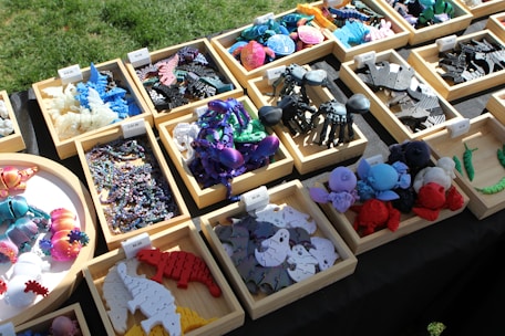 Various colorful 3D-printed creatures and objects are arranged in wooden trays on a table. The objects include dinosaurs, insects, and other shapes made from plastic. The scene is set outdoors with sunlight casting shadows, and each tray has a price label.
