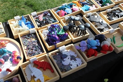 Various colorful 3D-printed creatures and objects are arranged in wooden trays on a table. The objects include dinosaurs, insects, and other shapes made from plastic. The scene is set outdoors with sunlight casting shadows, and each tray has a price label.