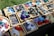Various colorful 3D-printed creatures and objects are arranged in wooden trays on a table. The objects include dinosaurs, insects, and other shapes made from plastic. The scene is set outdoors with sunlight casting shadows, and each tray has a price label.
