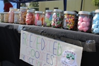 A table displays several jars filled with colorful treats, such as candies and snacks. Each jar has a screw-top lid, and there is a handwritten sign attached to the table that says 'FREEZE-DRIED GOODIES' and promotes the product, with a small logo of a company named 'Angry Armadillo'. The setting appears to be outdoors under a canopy.