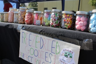 A table displays several jars filled with colorful treats, such as candies and snacks. Each jar has a screw-top lid, and there is a handwritten sign attached to the table that says 'FREEZE-DRIED GOODIES' and promotes the product, with a small logo of a company named 'Angry Armadillo'. The setting appears to be outdoors under a canopy.