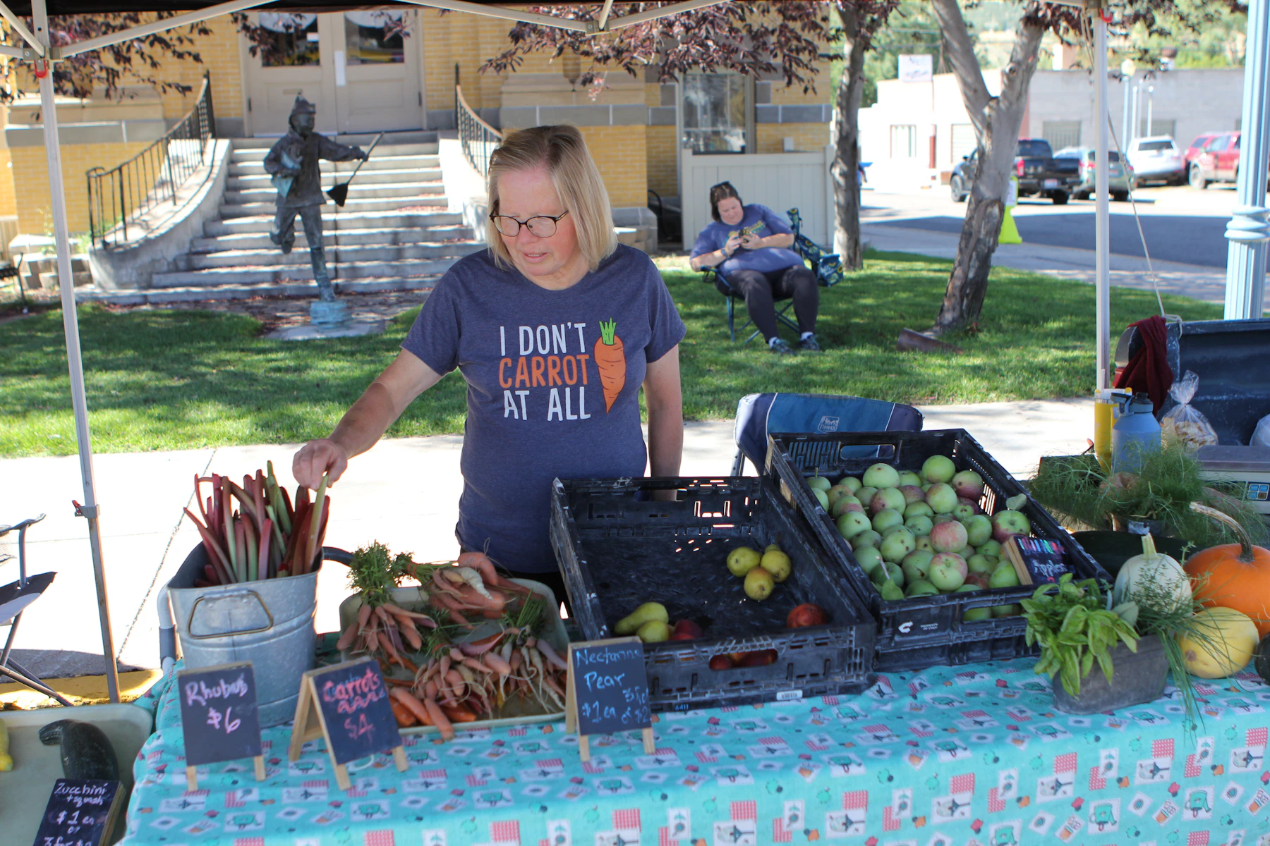 A grower at a farmers market stall under a canopy, with crates of apples, bundles of rhubarb and carrots, and chalkboard price signs