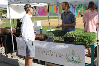 A small outdoor market or fair setting with two individuals engaged in conversation under a white tent. They stand near a table displaying an assortment of potted herbs and plants. The background features a colorful mural and a section of a park with a playground. The scene takes place on a sunny day.