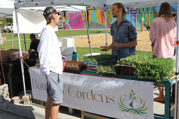 A small outdoor market or fair setting with two individuals engaged in conversation under a white tent. They stand near a table displaying an assortment of potted herbs and plants. The background features a colorful mural and a section of a park with a playground. The scene takes place on a sunny day.