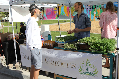 Farmers chatting and sharing stories with customers under green market tents on a sunny day.