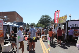 A lively outdoor market scene with people walking and conversing between food trucks parked on either side of a street. Brightly colored banners and flags, like 'Kettle Corn', are visible and add to the festive atmosphere. The setting is under clear blue sky with a distant view of trees and buildings.